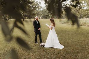Bride reading vows to groom outdoors under lush green trees during an intimate wedding ceremony.