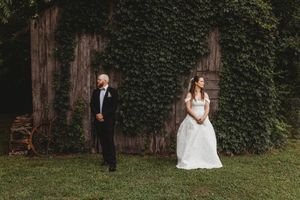 Bride in white gown and groom in tuxedo stand apart near ivy-covered barn wall on green grass.