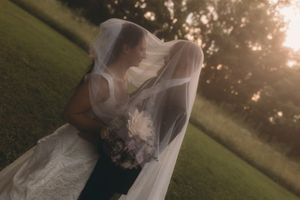 Bride and groom sharing a romantic moment under a flowing veil at golden hour in a lush green outdoor setting.