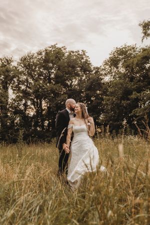 Romantic bride and groom embracing in golden meadow with tall grass and trees under cloudy sky.