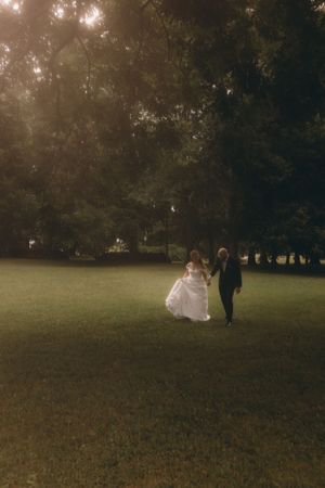 Bride and groom walking hand in hand across lush green lawn at dusk surrounded by tall trees.
