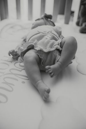 Black and white photo of a newborn baby lying in a crib wearing a knit romper, with chubby legs and tiny feet visible.