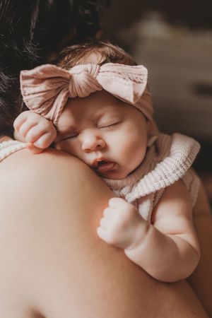 Newborn baby girl wearing a pink bow headband sleeping peacefully on mother's shoulder in soft warm light.