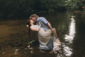 Couple sharing a romantic kiss while standing in a shallow forest creek, surrounded by lush green trees.