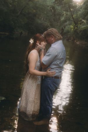 Romantic couple embracing in a sunlit forest creek, woman in floral dress with flower in hair, man in blue shirt and jeans.