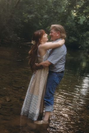 Couple embracing romantically in a shallow creek surrounded by lush green trees in a dreamy outdoor setting.