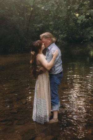 Couple sharing a romantic kiss while standing in a shallow forest creek, woman in lace dress, man in jeans.