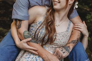 Couple embracing outdoors, woman in floral dress with tattoos smiling, man hugging her from behind.