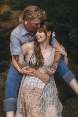 A couple embraces outdoors, woman in floral dress with flower in hair, man kissing her temple near lush greenery.