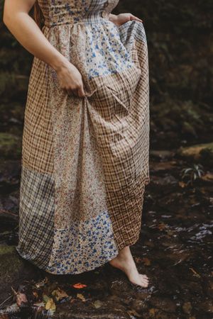 Woman in patchwork floral and plaid maxi dress lifting hem while standing barefoot in a forest stream.