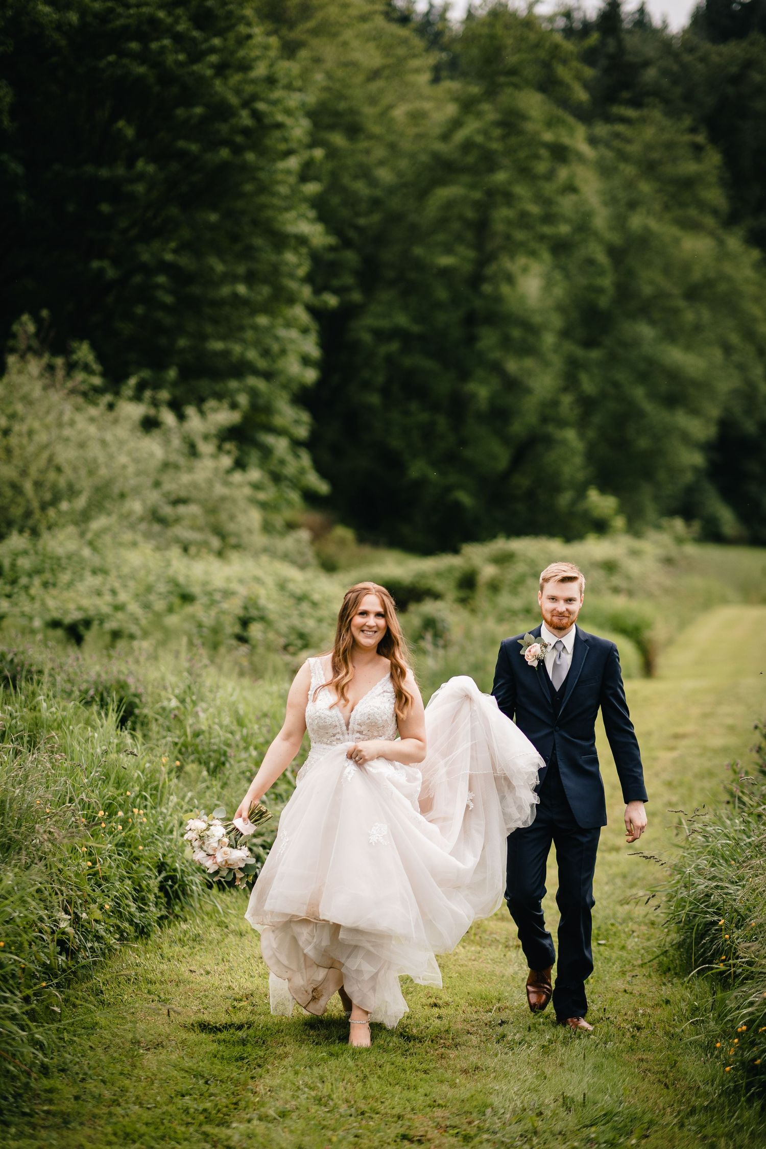 A couple walks along a grassy path through lush greenery, with the bride wearing a flowing white gown and groom in dark suit.