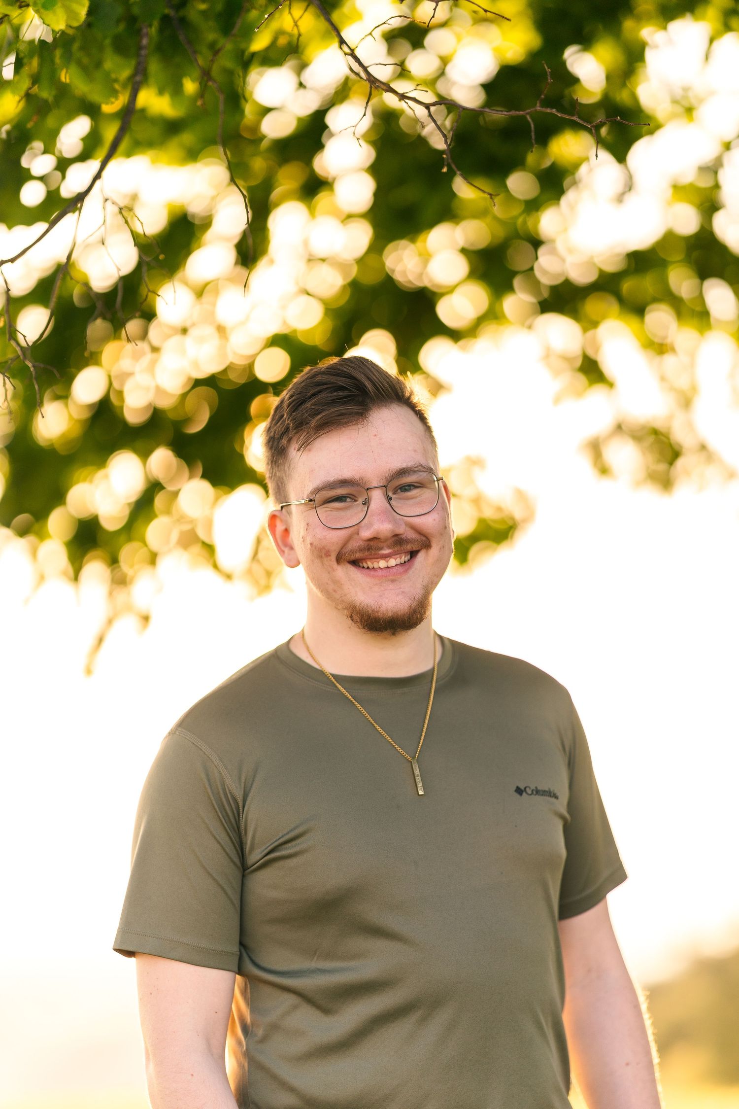 A person in a green t-shirt stands under sunlit tree branches.