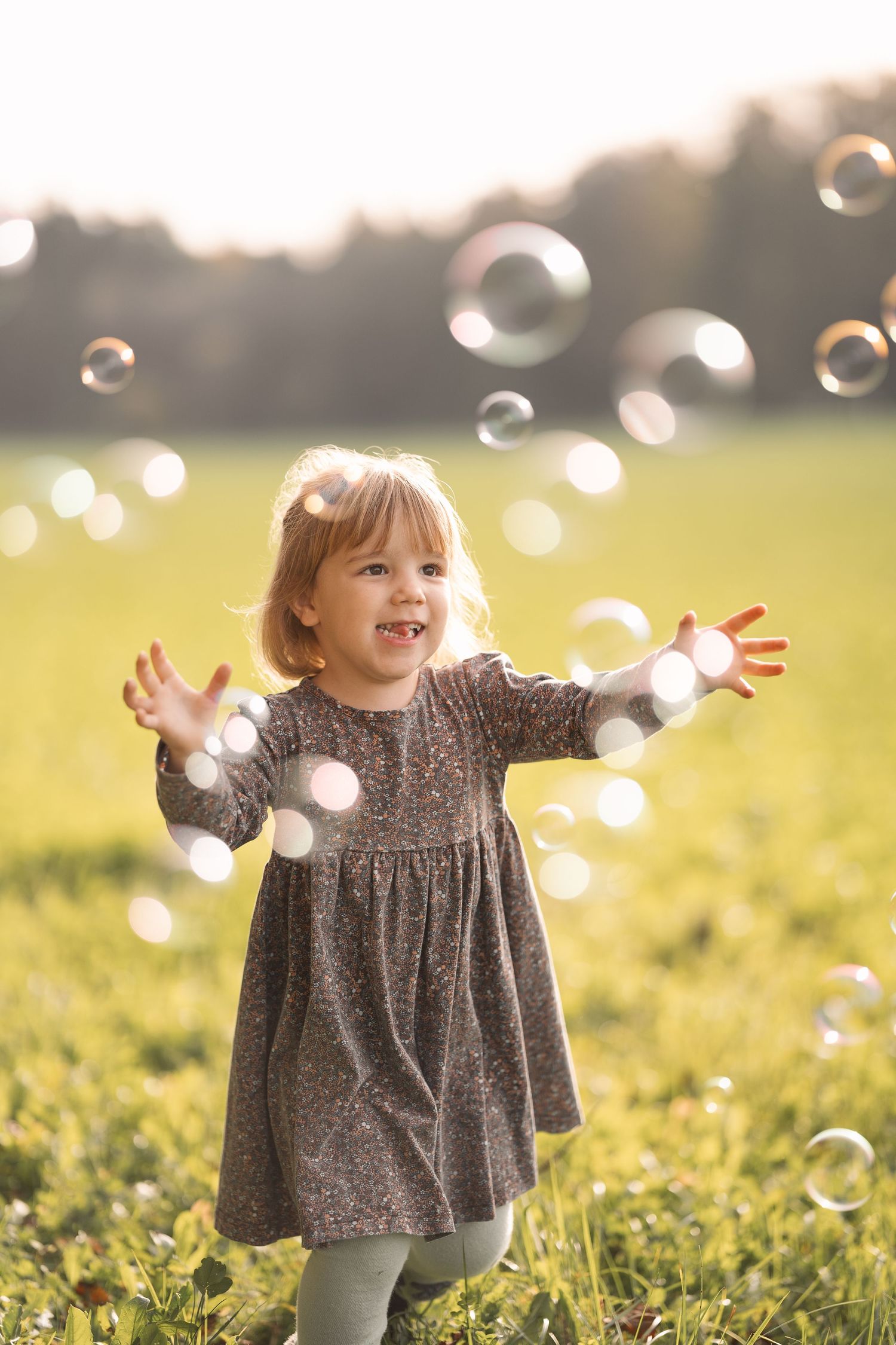 A child plays with bubbles in a sunny field wearing a gray dress.