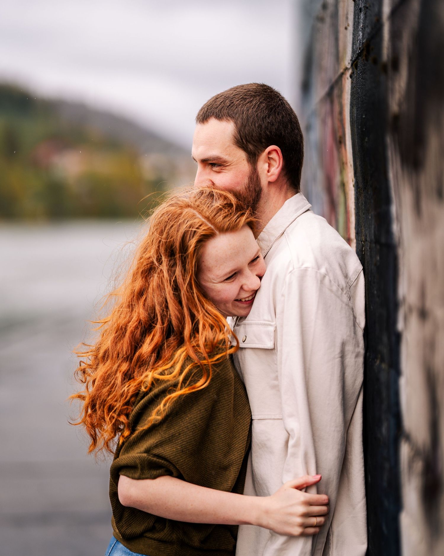 A tender embrace captured against a lakeside backdrop on an overcast day.