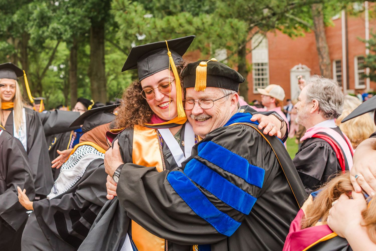 Graduates in black gowns with orange stoles and blue regalia celebrating together during a commencement ceremony.