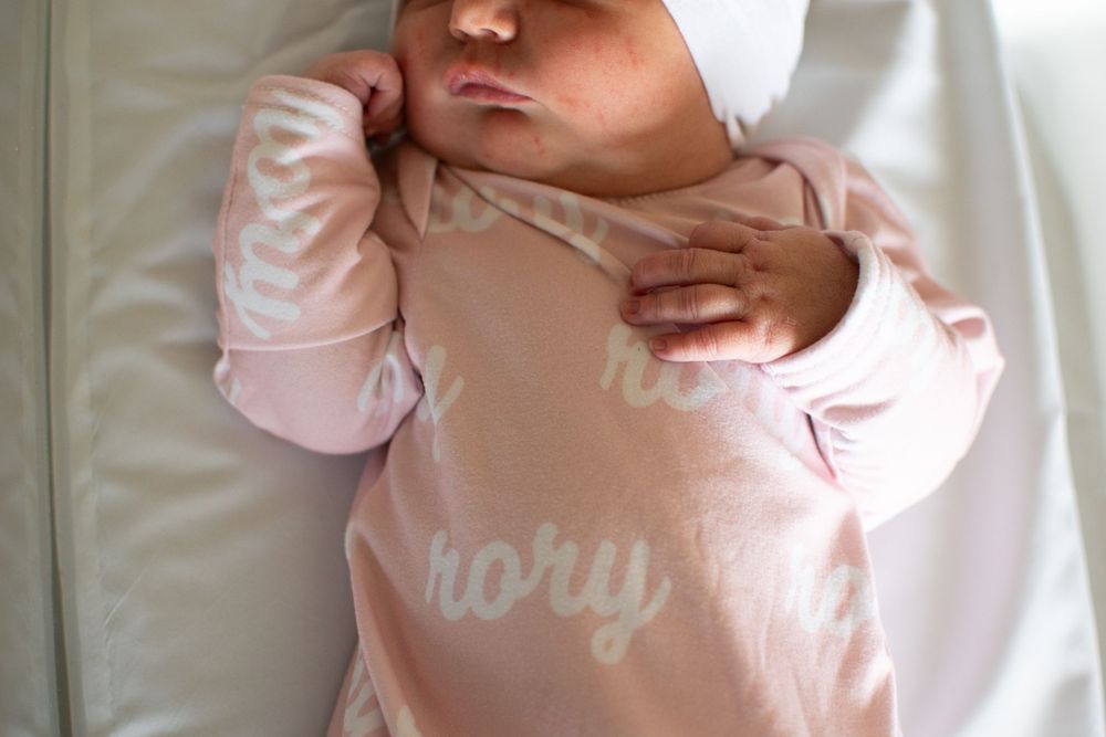 Baby wearing pink onesie with 'story' text lying on white bedding.