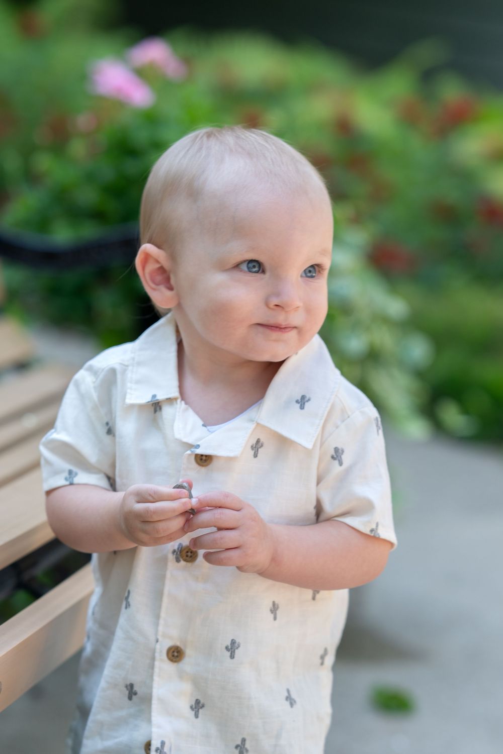 Young child wearing a white patterned button-down shirt smiles while standing outdoors in a garden setting.