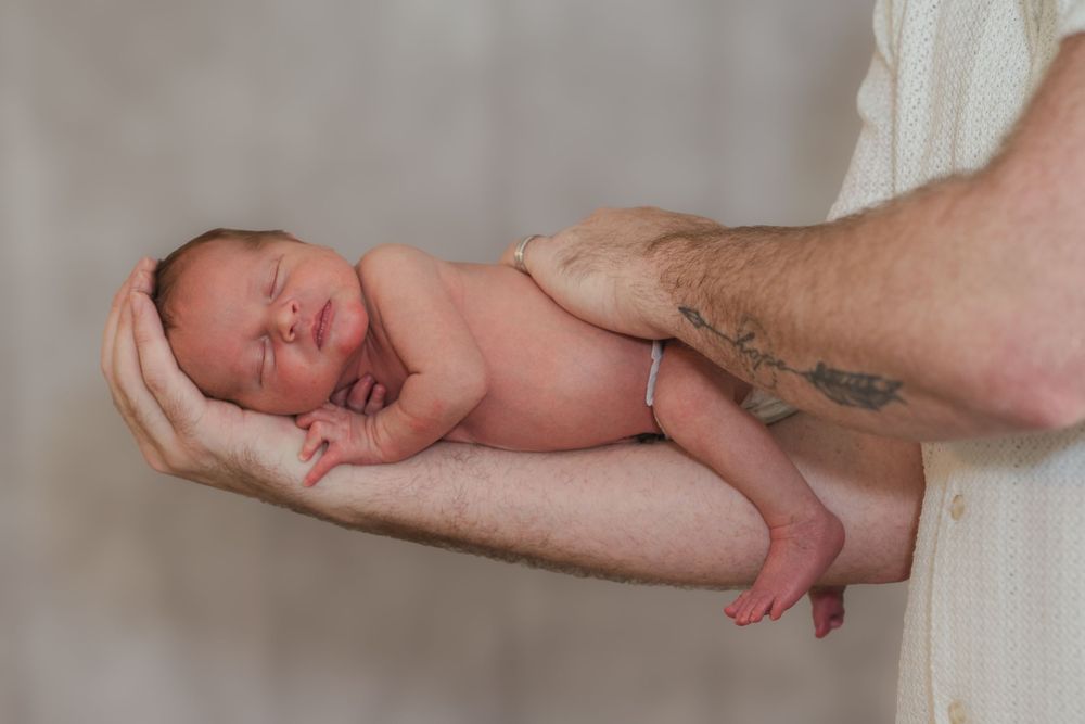 Newborn baby lying on white fabric while adult hands gently support and position them for a photo session.