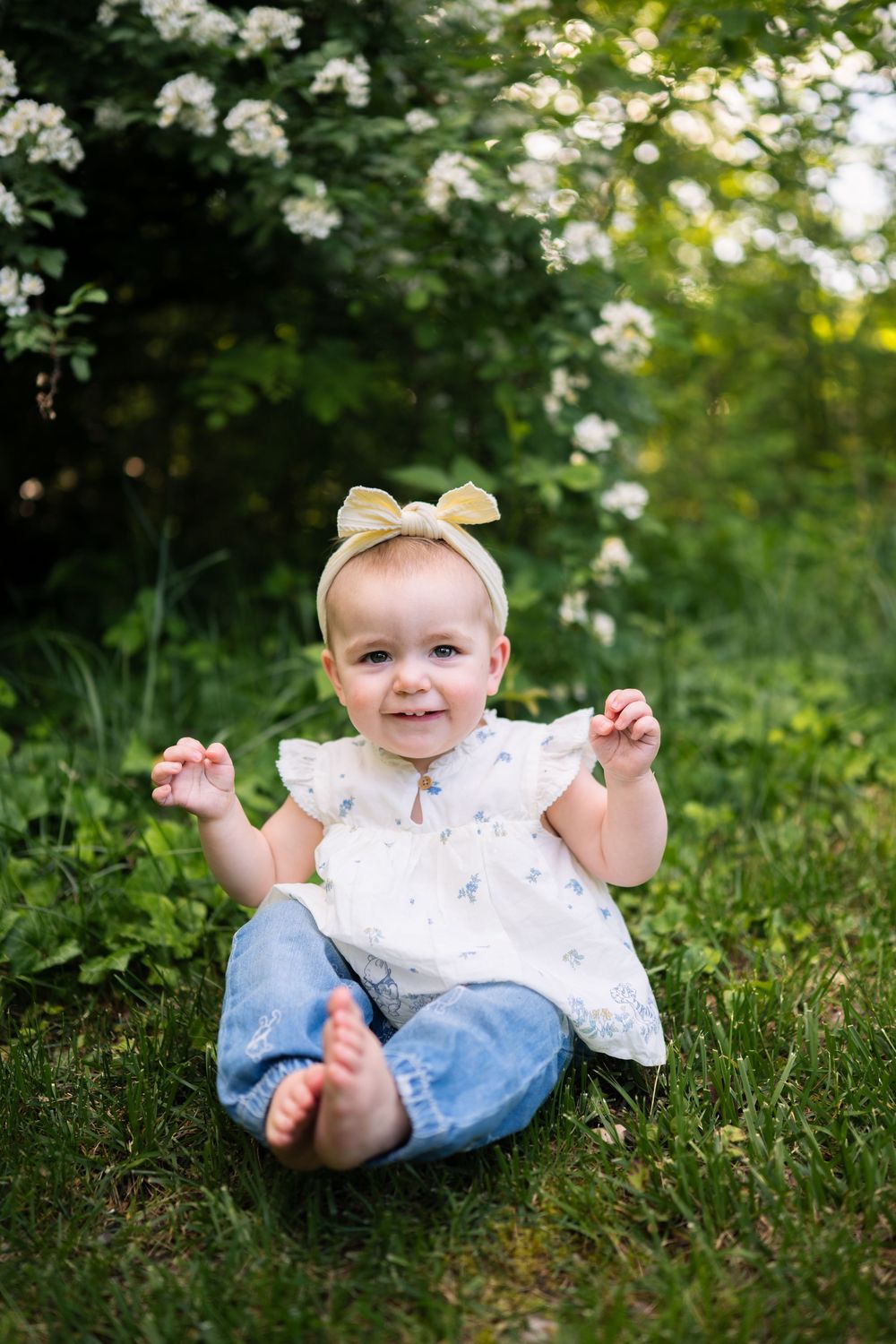 Smiling baby girl in floral top, jeans, and yellow bow headband sitting barefoot on green grass outdoors.