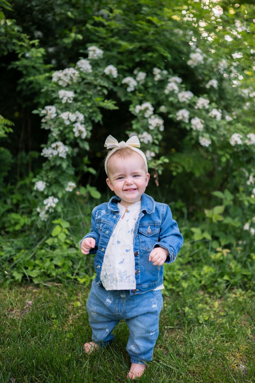 Smiling baby girl in denim jacket and jeans with bow headband standing on grass near white flowering bushes.