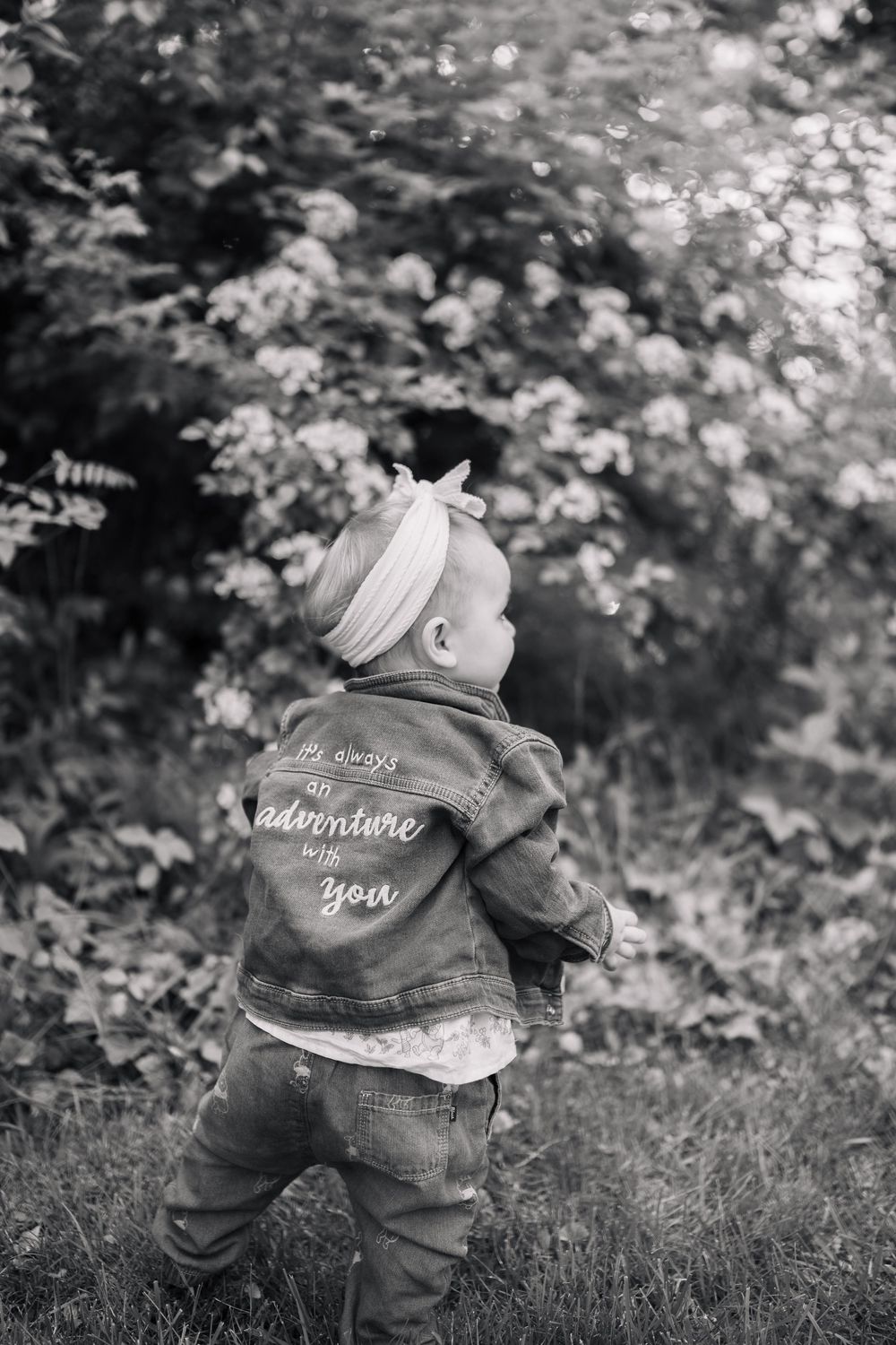 Black and white photo of a baby girl in a denim jacket with text, wearing a headband, exploring outdoors.
