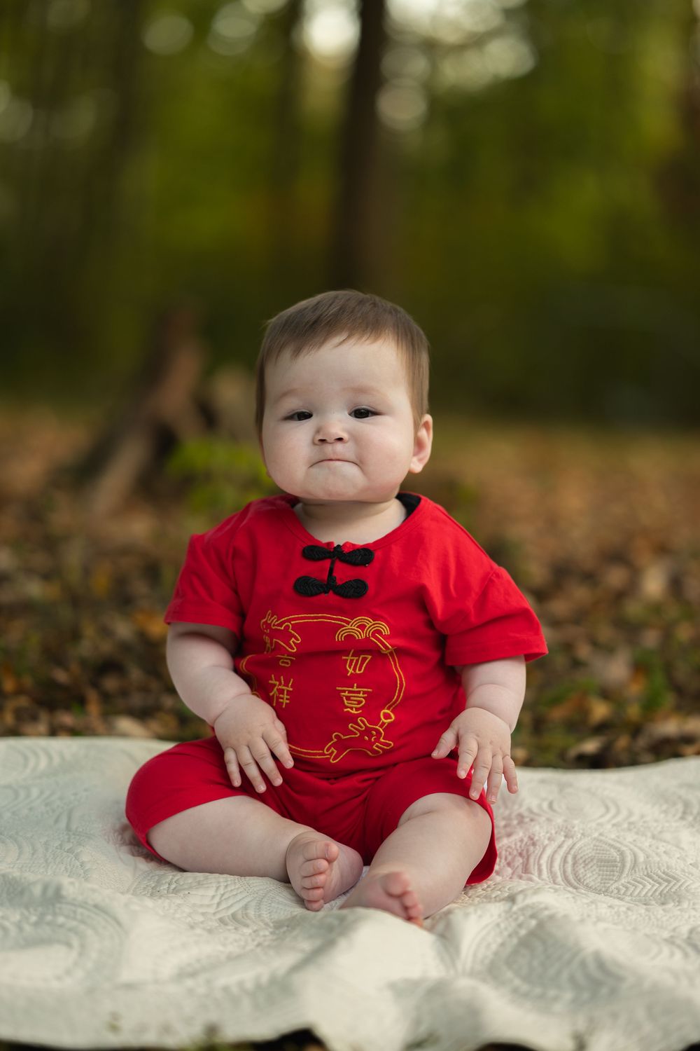 A baby wearing a festive red outfit sits on a white blanket outdoors with autumn foliage in the background.