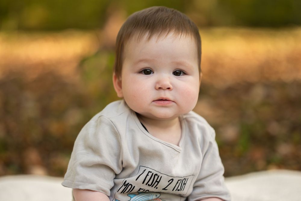 A baby wearing a light colored onesie sits outdoors on a sunny autumn day with fallen leaves in the background.