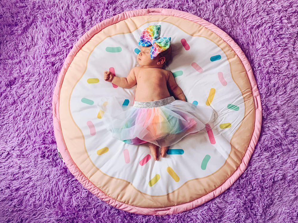 Baby wearing colorful tutu lying on a donut-shaped play mat with pastel decorations against purple background.