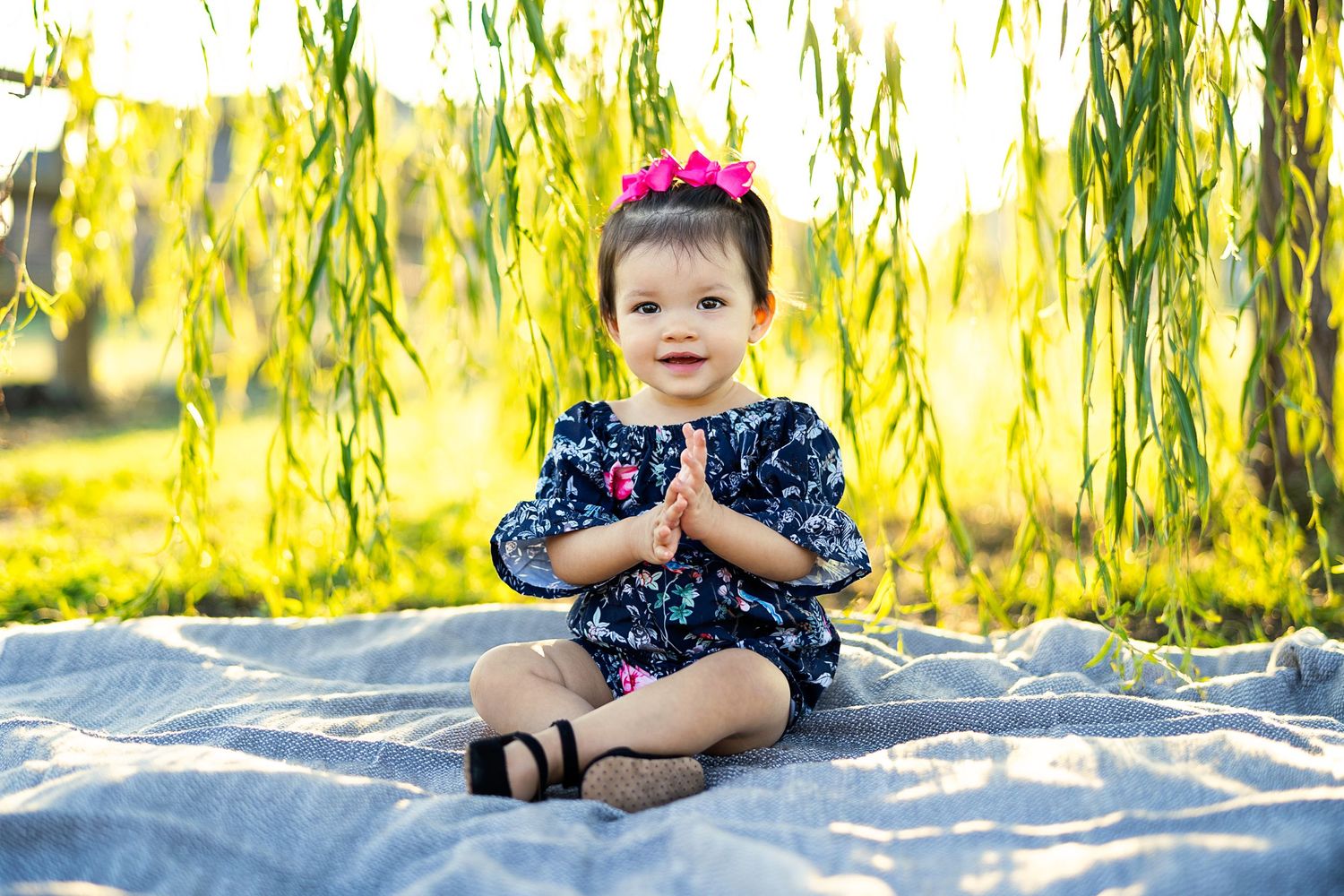 Baby in floral dress sitting on blanket under willow tree in sunlit garden.