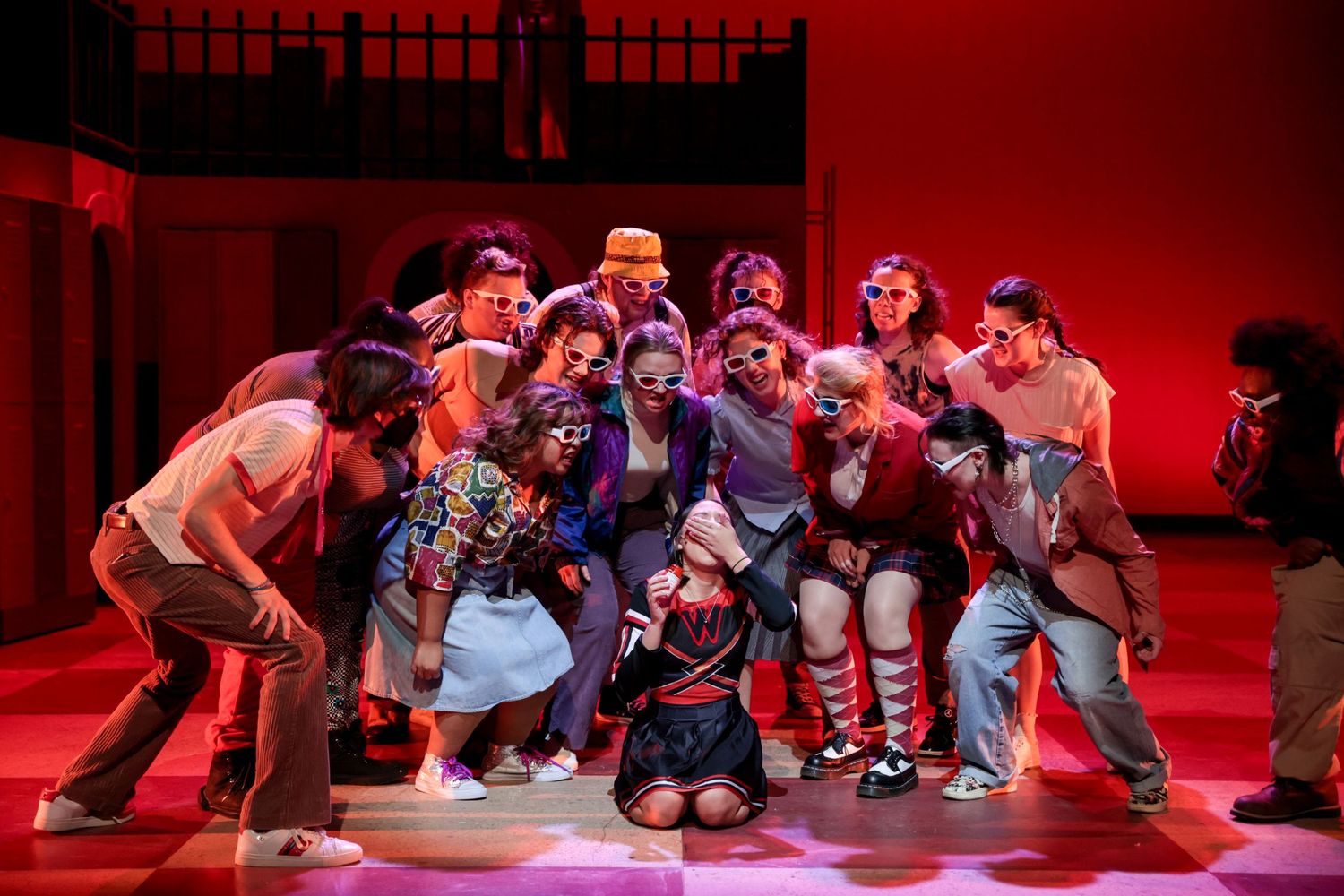 Energetic theater ensemble performs together under red lighting on a multi-level stage set.