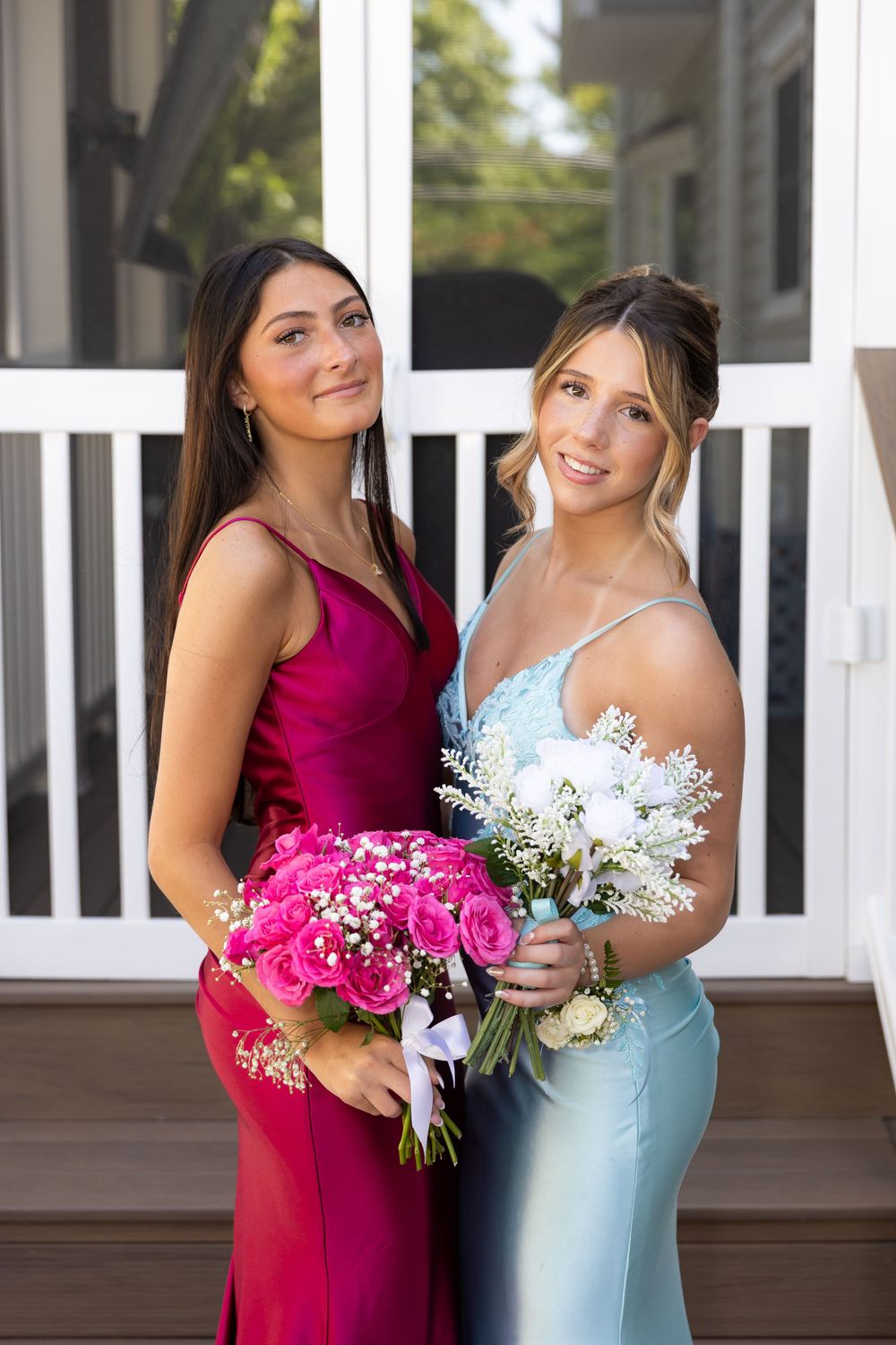 Two young women in formal prom gowns pose holding pink and white floral bouquets outdoors.