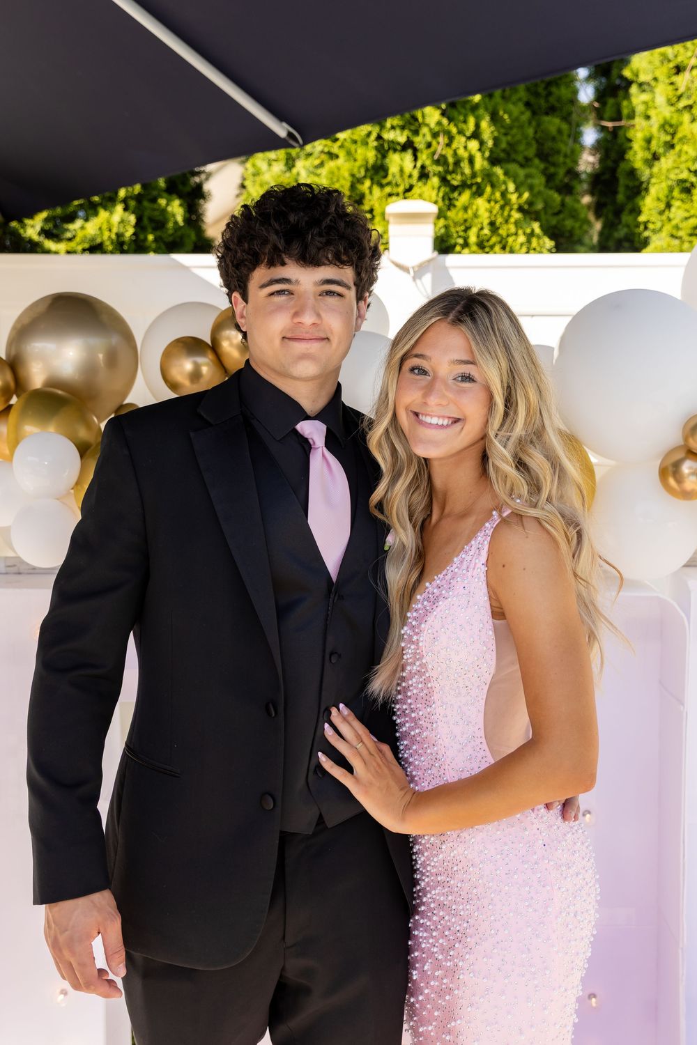 Smiling teenage prom couple pose together outdoors with gold and white balloon backdrop.