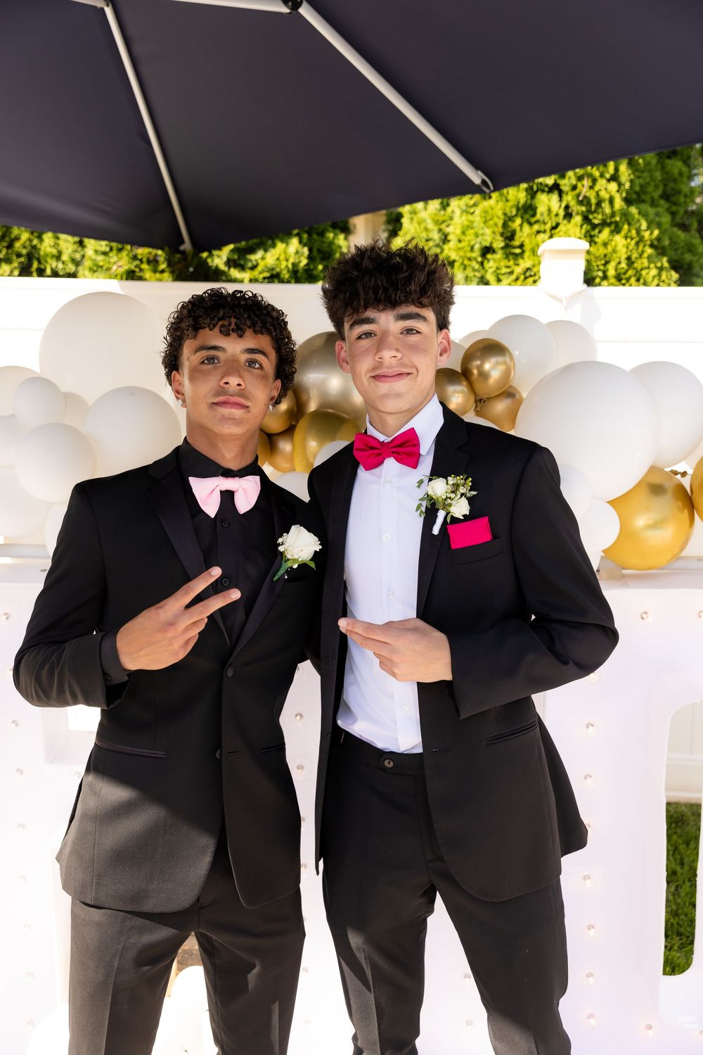 Two teenage boys in black tuxedos with bow ties and boutonnieres pose at a prom photo booth with gold and white balloons.