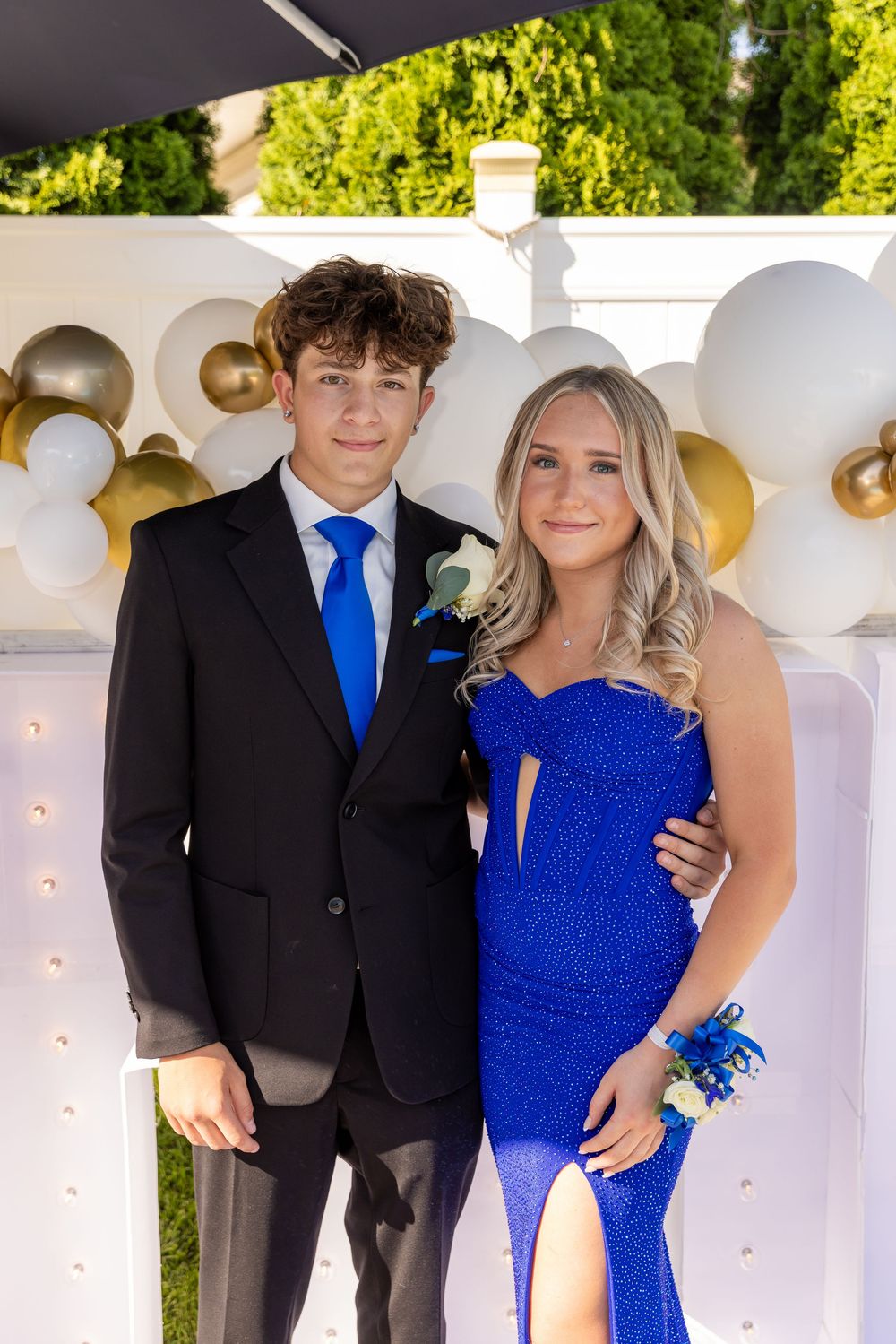 A teenage couple in prom attire, black tuxedo and royal blue gown, pose together in front of a balloon backdrop.