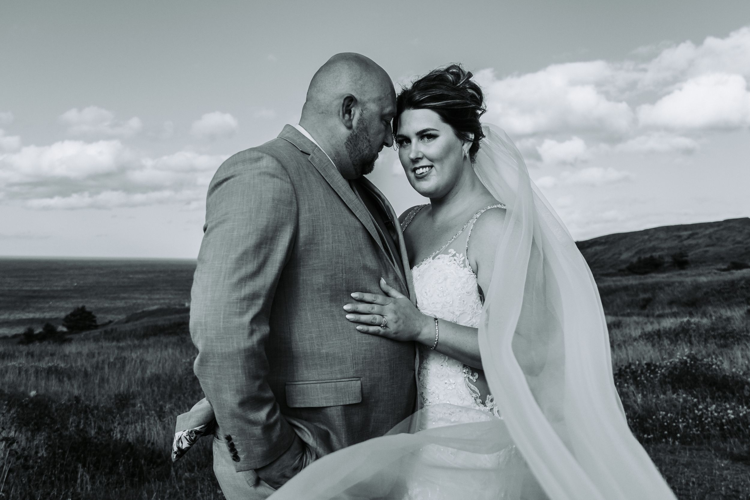 A romantic black and white wedding photo captured against a scenic rural hillside landscape background.