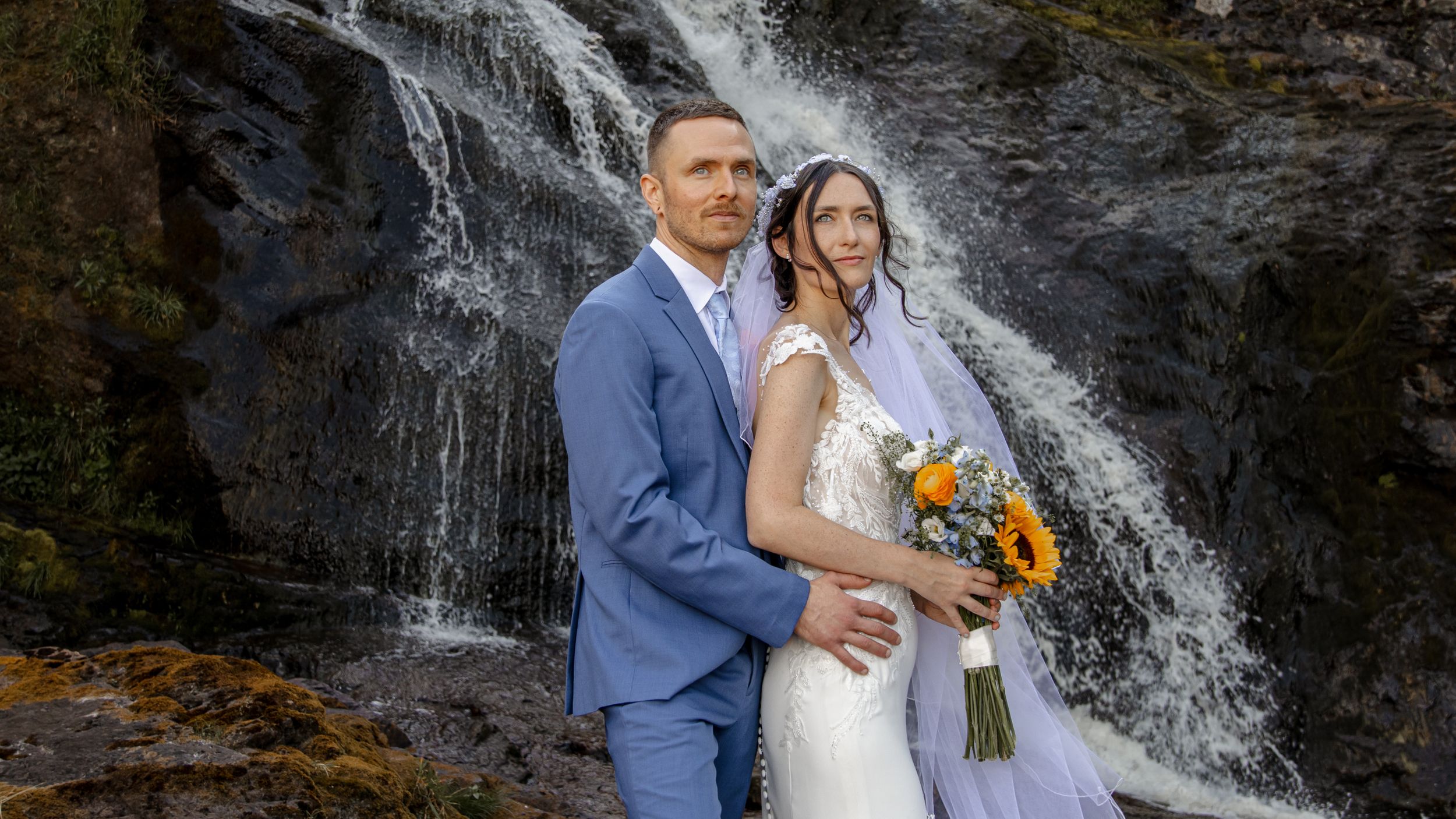 A couple poses in formal wedding attire in front of a cascading waterfall with a sunflower bouquet.