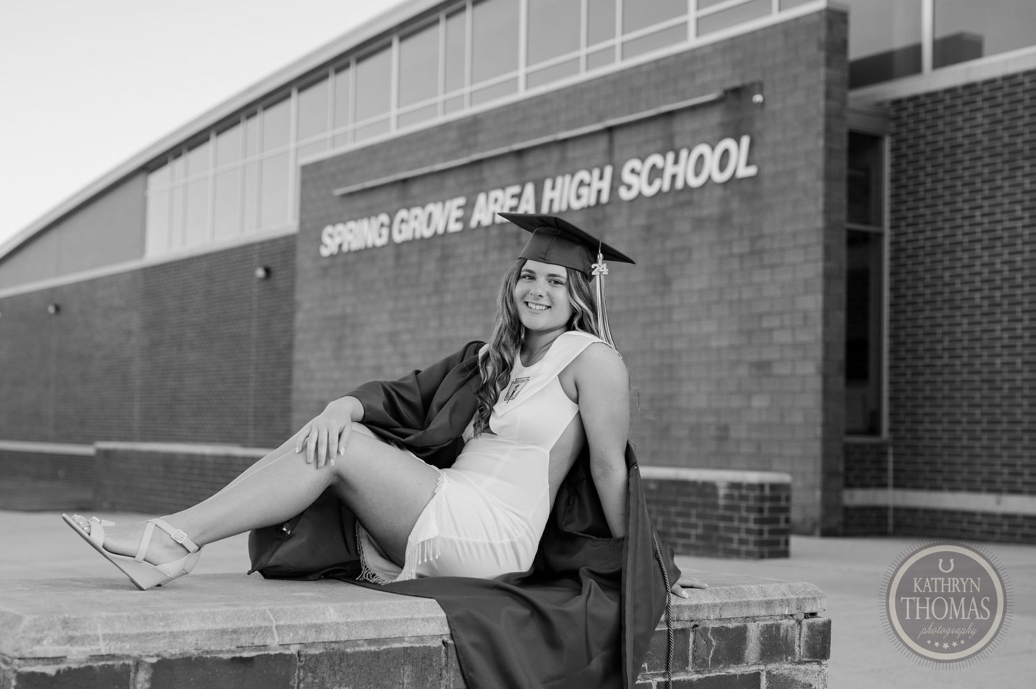 High school graduate in cap and gown sits casually outside school building.