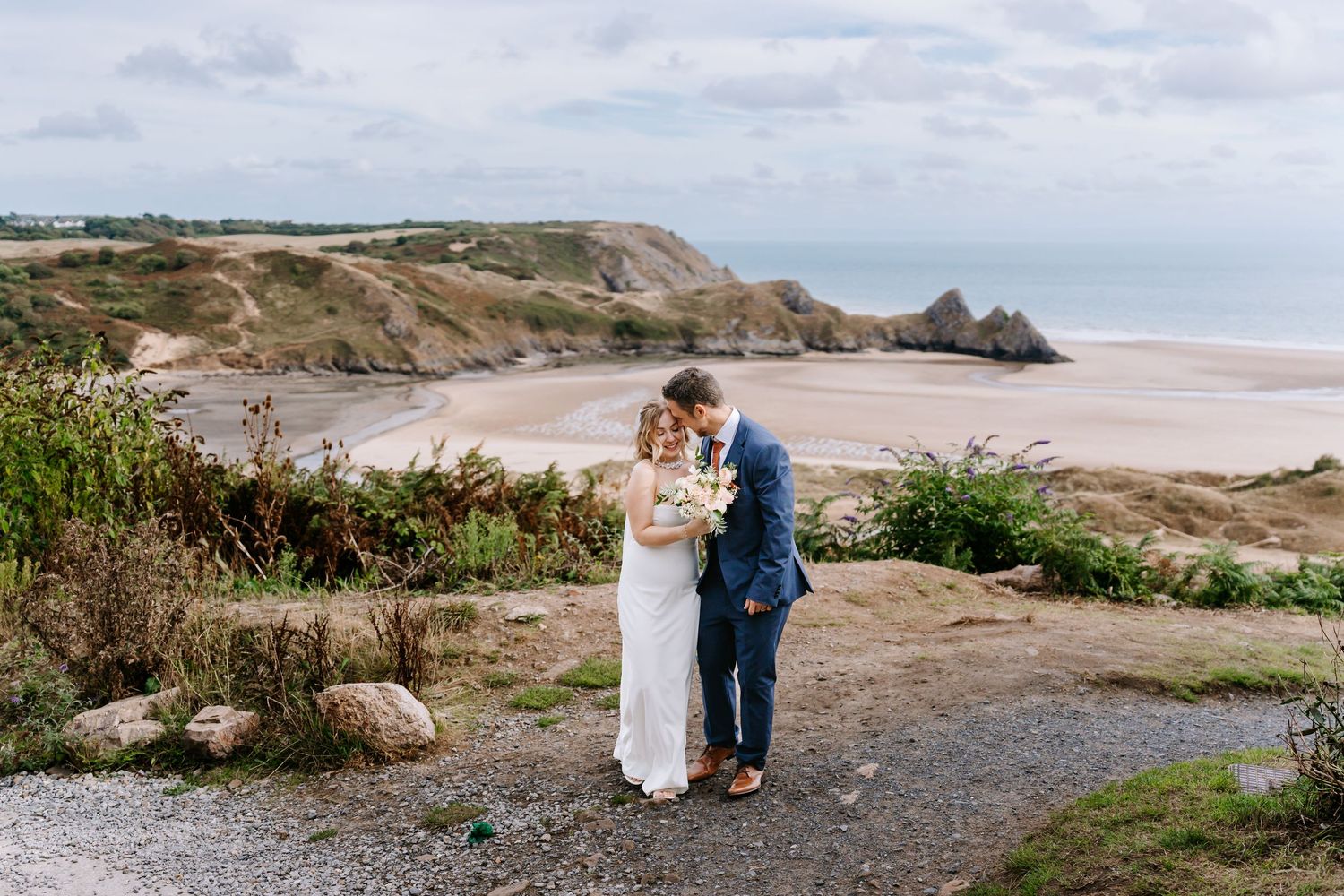 A couple in wedding attire walking on a beach at sunset with ocean views.