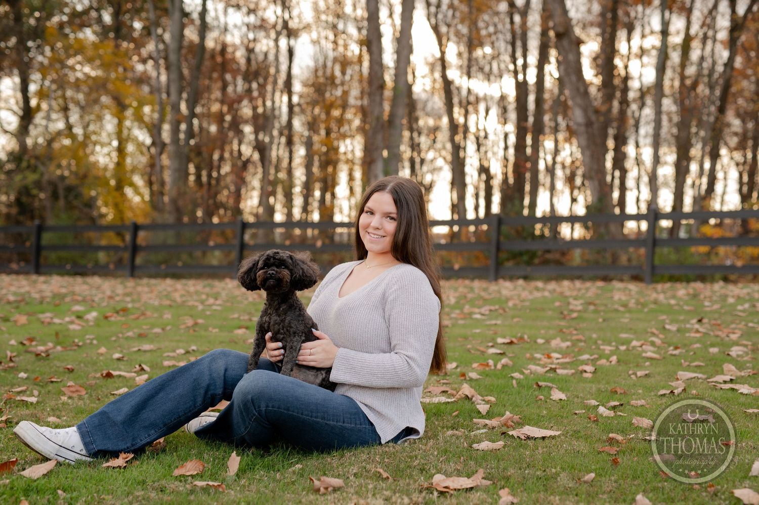 Person sits on grass with small black dog during autumn photoshoot with trees in background.