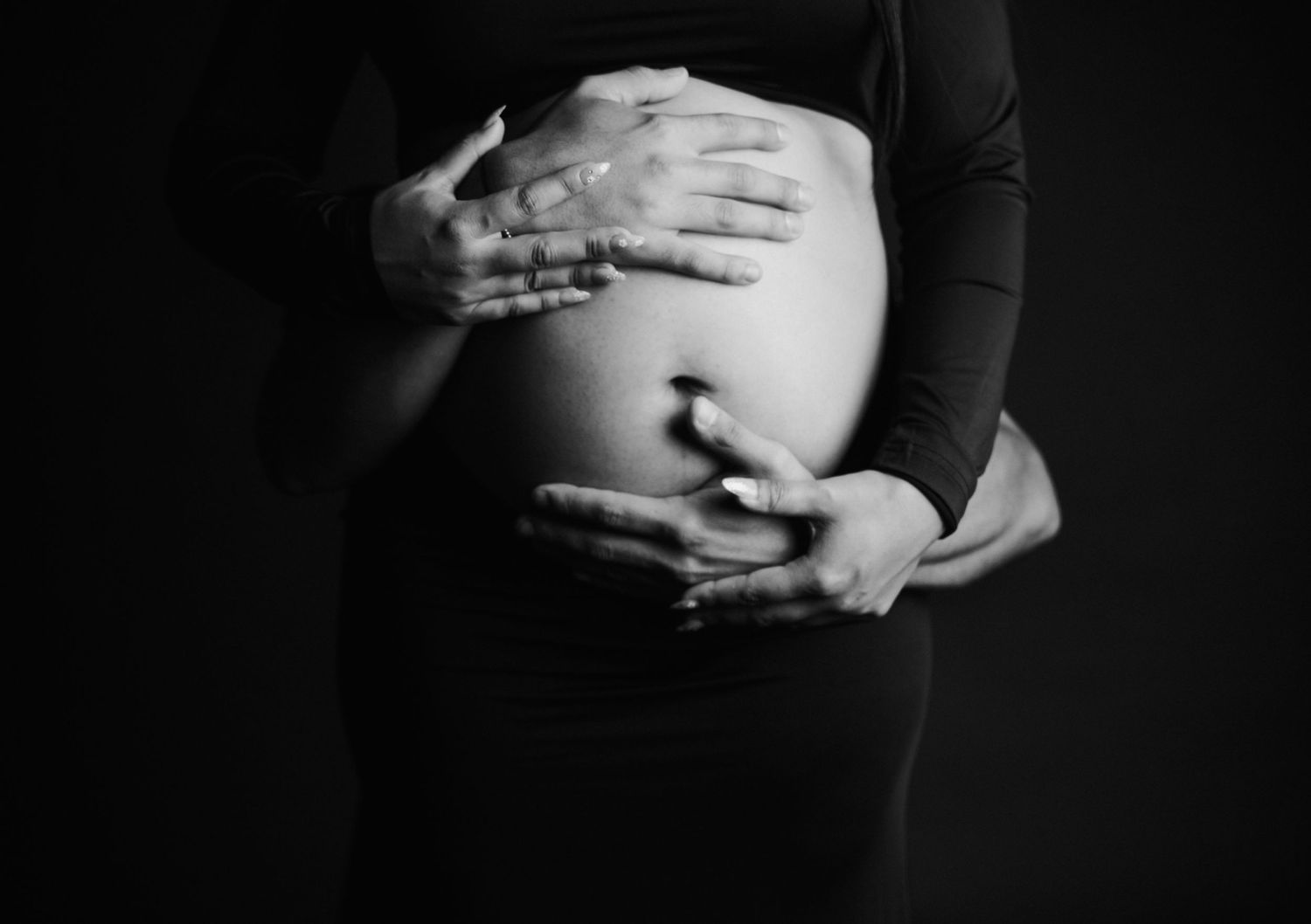 Black and white photo of hands tenderly cradling a pregnant belly against dark background.