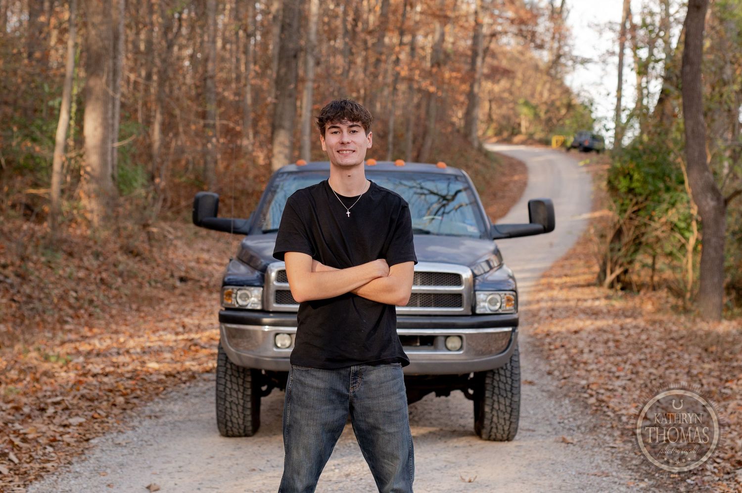 Person standing with crossed arms in front of silver Dodge Ram truck on autumn forest path.