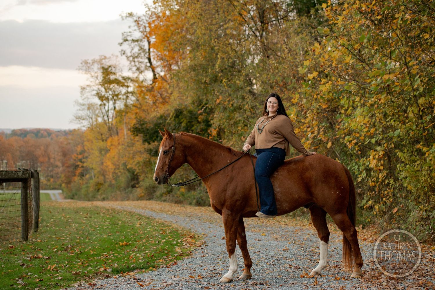 A rider sits atop a brown and white horse on a scenic fall trail with autumn foliage in the background.