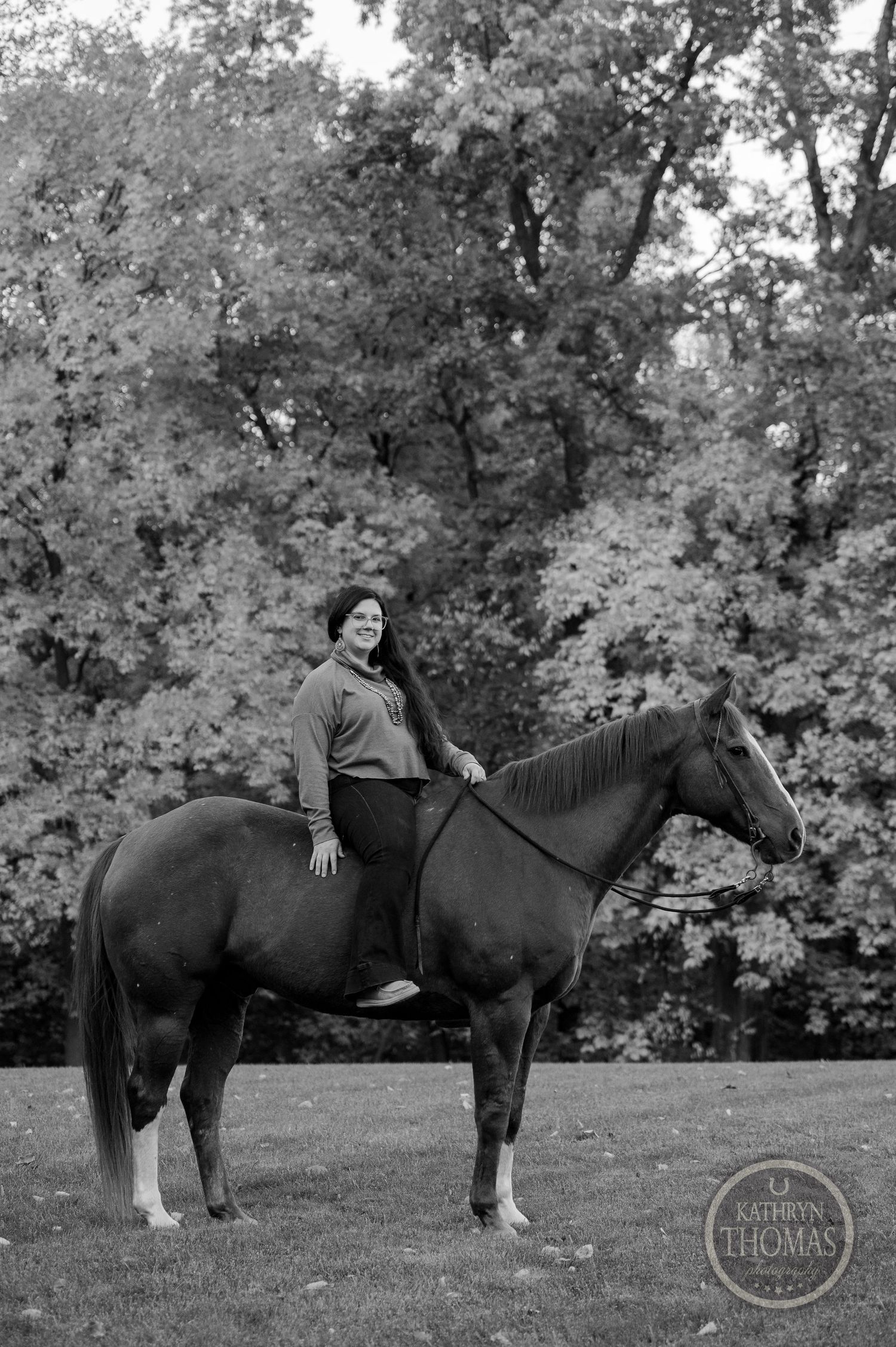 A black and white photo captures a horse and rider posing elegantly among autumn trees.