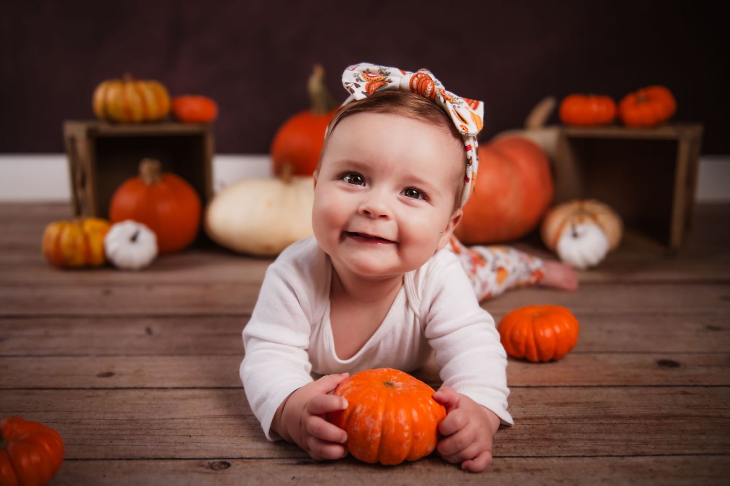Small baby in autumn setting with orange pumpkins and wooden crates on rustic floor.