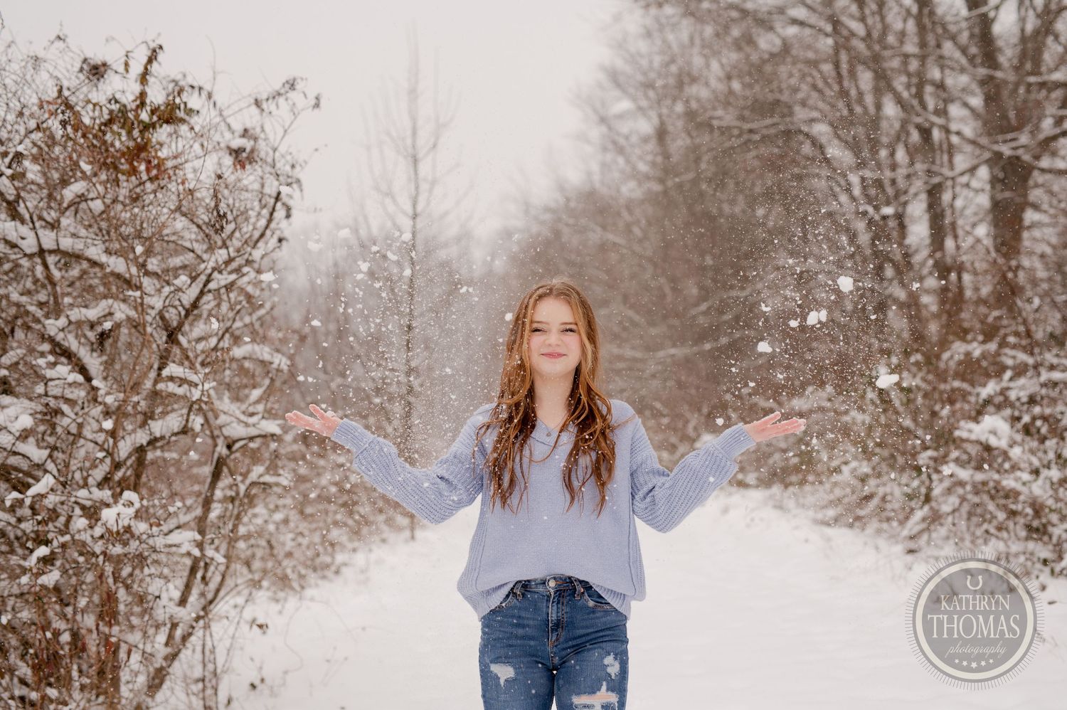 Someone spreads their arms joyfully on a snow-covered path wearing a light blue sweater.