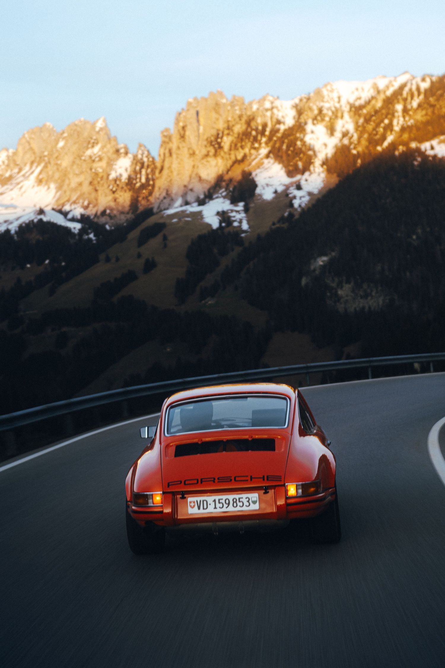 Classic orange Porsche driving on mountain road with snow-capped peaks at sunset.