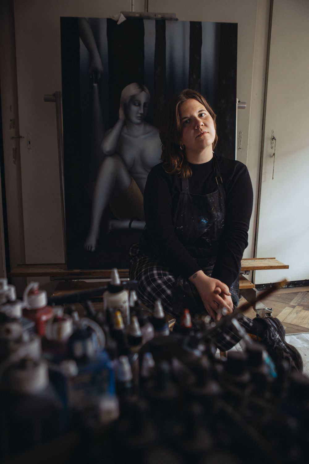 Artist in dark clothing surrounded by art supplies with a moody black and white portrait in the background.