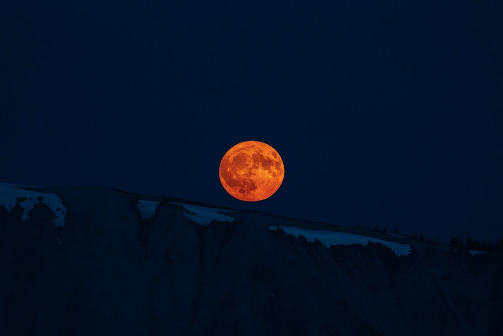 Blood orange full moon rising over mountain silhouettes against dark night sky.