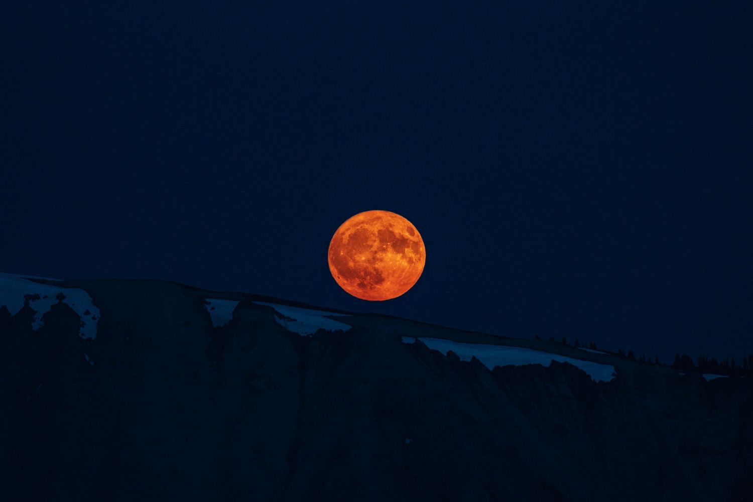 Blood orange full moon rising over mountain silhouettes against dark night sky.