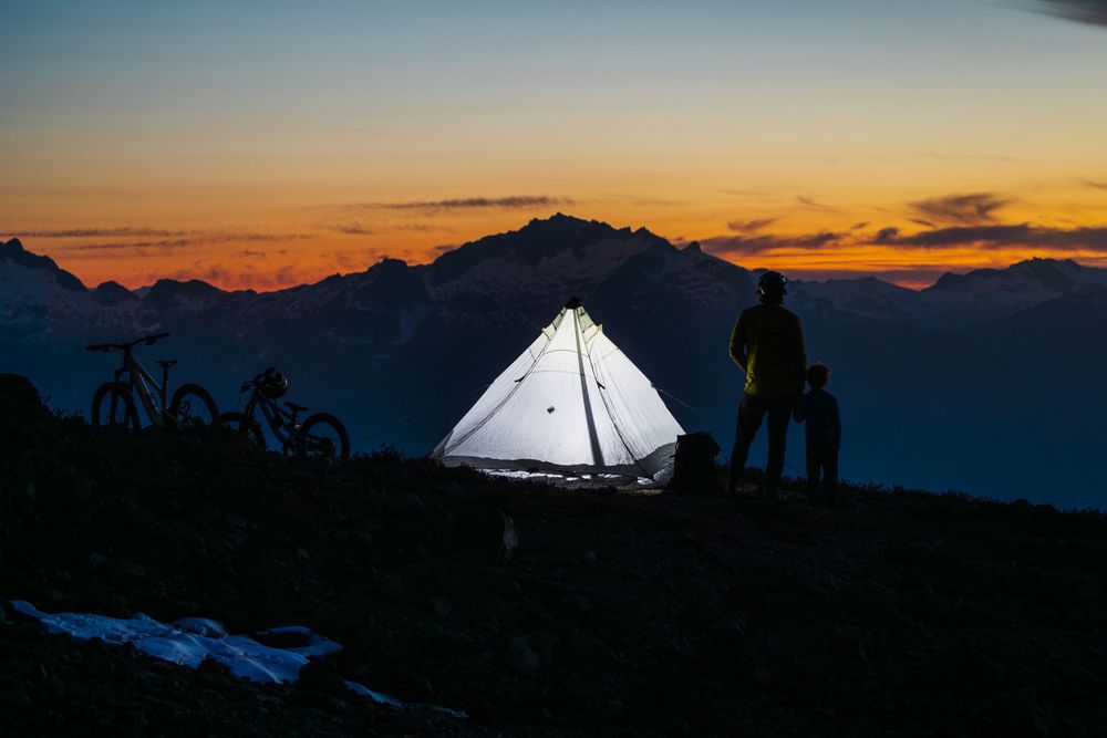 Illuminated tent glows against mountain silhouette during colorful sunset with bikes and figures.